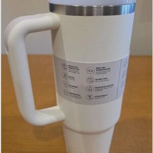 May include: A white stainless steel tumbler with a large side handle, a clear lid, and a straw. A paper band around the center lists features including recycled 18/8 stainless steel, double-wall vacuum insulation, BPA-free, and dishwasher safe.