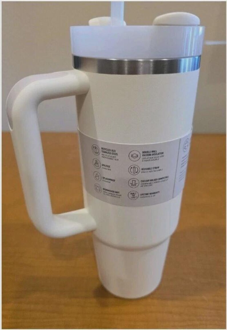 May include: A white stainless steel tumbler with a large side handle, a clear lid, and a straw. A paper band around the center lists features including recycled 18/8 stainless steel, double-wall vacuum insulation, BPA-free, and dishwasher safe.