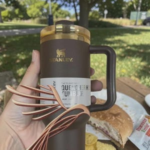 Puede incluir: Un vaso Stanley Quencher H2.0 marr&oacute;n con tapa y asa doradas. El vaso tiene el logotipo de Stanley y el texto "QUENCHER H2.0 TUMBLER" en la etiqueta. El vaso tiene una capacidad de 1,18 litros.