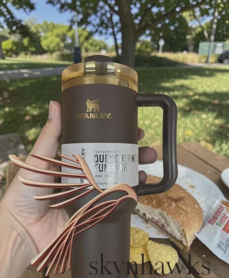 Puede incluir: Un vaso Stanley Quencher H2.0 marr&oacute;n con tapa y asa doradas. El vaso tiene el logotipo de Stanley y el texto "QUENCHER H2.0 TUMBLER" en la etiqueta. El vaso tiene una capacidad de 1,18 litros.
