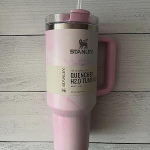 K&ouml;nnte beinhalten: Ein hellrosa Stanley Quencher H2.0 Becher mit marmoriertem Muster, einem Griff und einem durchsichtigen Strohhalm. Das Produktetikett zeigt den Text Stanley, The Flowstate, Quencher H2.0 Tumbler, 40 oz, 1.18 L und All Day Hydration.