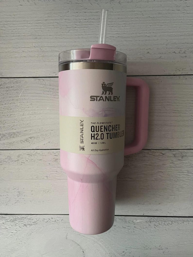 K&ouml;nnte beinhalten: Ein hellrosa Stanley Quencher H2.0 Becher mit marmoriertem Muster, einem Griff und einem durchsichtigen Strohhalm. Das Produktetikett zeigt den Text Stanley, The Flowstate, Quencher H2.0 Tumbler, 40 oz, 1.18 L und All Day Hydration.