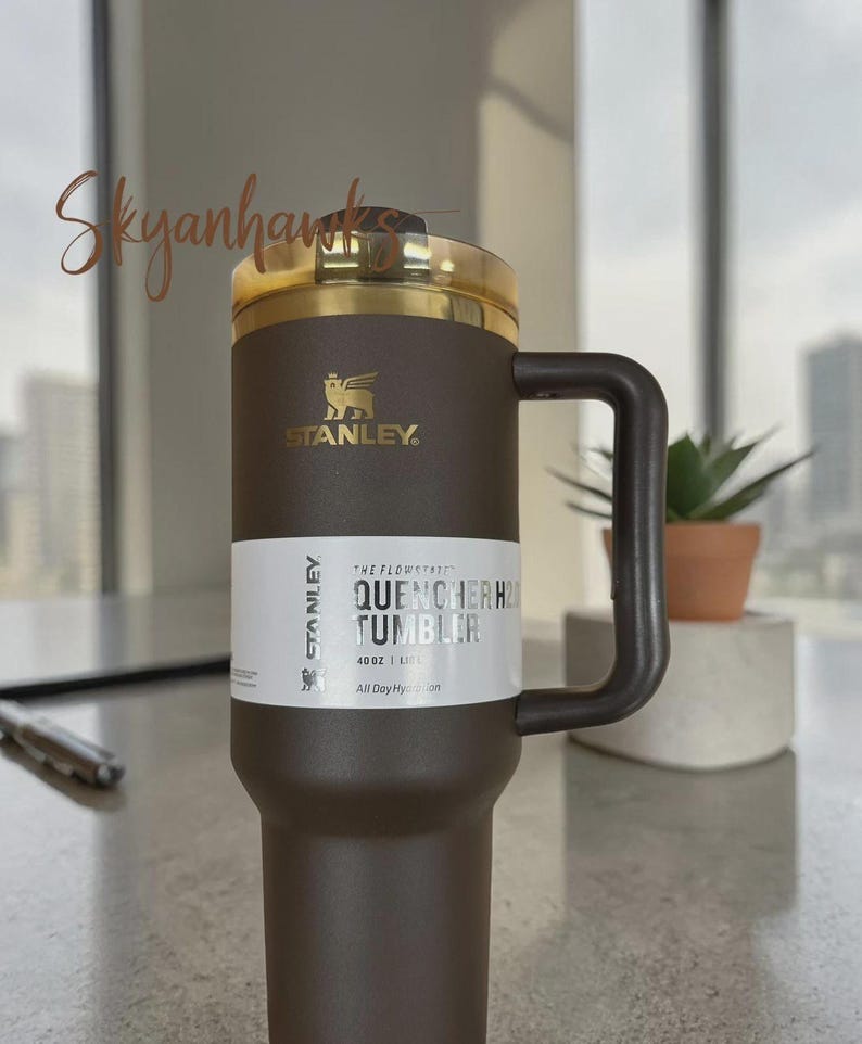 Puede incluir: Un vaso Stanley Quencher H2.0 de color marr&oacute;n oscuro con una capacidad de 1,18 L. El vaso cuenta con un asa a juego, una tapa de tono dorado y una etiqueta blanca con el texto The Flowstate Quencher H2.0 Tumbler 40 oz 1.18 L All Day Hydration.