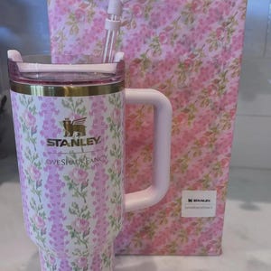 K&ouml;nnte beinhalten: Ein Stanley-Becher mit floralem Muster, Griff, Strohhalm und Deckel, zusammen mit einer passenden Geschenkbox. Der Becher hat ein rosa-wei&szlig;es Blumenmuster mit goldenen Akzenten und das Logo "Stanley Loveshackfancy".