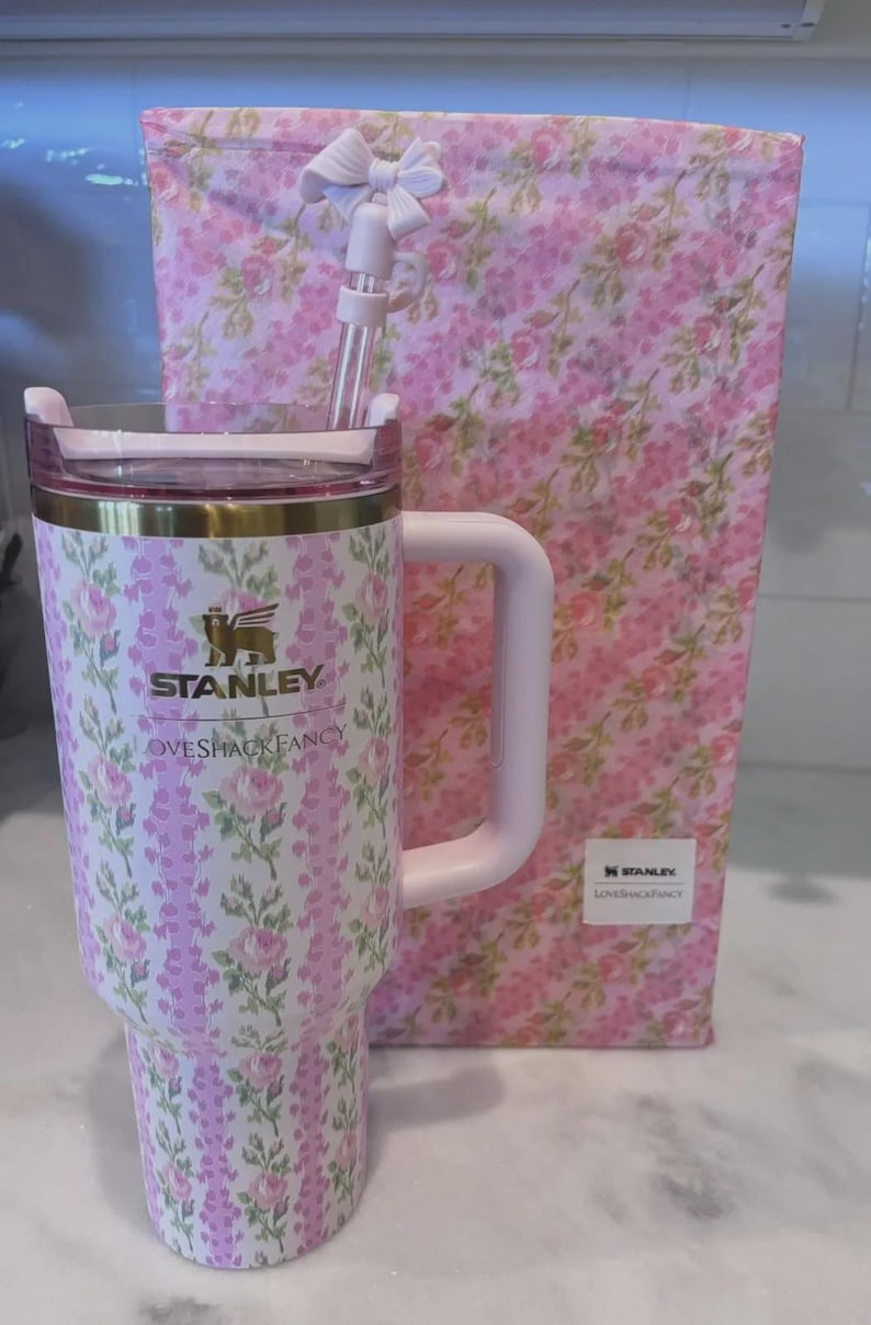 K&ouml;nnte beinhalten: Ein Stanley-Becher mit floralem Muster, Griff, Strohhalm und Deckel, zusammen mit einer passenden Geschenkbox. Der Becher hat ein rosa-wei&szlig;es Blumenmuster mit goldenen Akzenten und das Logo "Stanley Loveshackfancy".