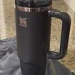 May include: A black insulated tumbler with a handle and a lid. The tumbler has a stainless steel rim and a logo on the side. The tumbler is sitting on a gray cloth.
