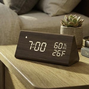 Puede incluir: Un reloj despertador digital de madera que muestra las 7:00, 60% HR y 26F. El reloj es triangular y está sobre una mesita de noche de madera junto a una cama con almohadas y una lámpara. La habitación tiene una paleta de colores neutros.