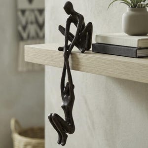 Pode incluir: Uma escultura em bronze de duas figuras, uma sentada e outra pendurada, estendendo as mãos uma para a outra. A escultura está numa prateleira de madeira clara, com uma pequena planta num vaso cinzento e livros.