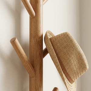 May include: A light brown wooden coat rack with a central vertical post and multiple angled pegs. A tan woven straw fedora hat with a brown ribbon band is hanging from one of the wooden pegs, showcasing the detailed texture of the hat's weave.