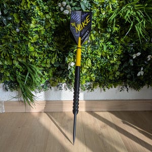 Op de afbeelding: Een zwarte en gele dartpijl met een zwarte schacht en een punt. De dartpijl heeft een zwarte en blauwe flight met de tekst "THE NUKE TARGET". Hij staat tegen een groene plantenmuur en een houten vloer.