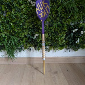 Op de afbeelding: Een dartpijl met een paarse flight voorzien van gele penseelstreken met de tekst NUK, een paarse shaft en een zilverkleurige barrel met een geribbelde grip. De pijl eindigt in een goudkleurige punt en staat rechtop op een houten ondergrond.