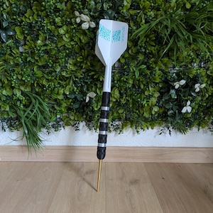 Puede incluir: Un dardo con una pluma blanca con un diseño turquesa. El dardo tiene un agarre negro y plateado y una punta dorada. Se encuentra sobre una superficie de madera, con una pared de plantas verdes al fondo.