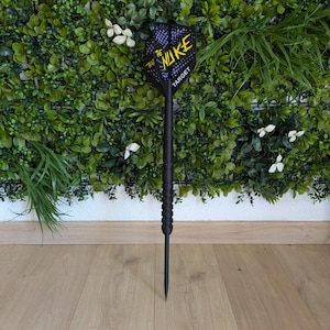 May include: A black dart with a patterned flight displaying the text "THE NUKE TARGET" in yellow and white. The dart is set against a backdrop of green foliage and white flowers, with a wooden floor.