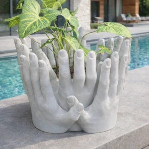 May include: A distinctive grey concrete planter, sculpted in the form of hands, holding a plant with large green and yellow leaves. The planter is placed outside near a swimming pool and a modern house.