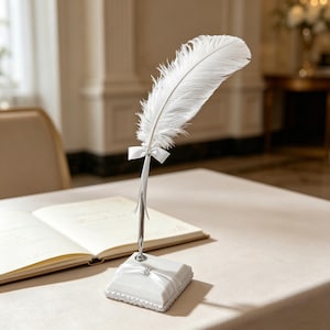 May include: A white feather quill pen with a silver-colored metal base and a small white bow. The pen rests on a white square base with a ribbon and a small jewel. An open book is in the background.