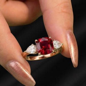 May include: A gold ring featuring a large, square-cut, deep red gemstone flanked by two pear-shaped, clear gemstones. The ring is held against a black background.