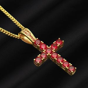 Könnte beinhalten: Eine goldfarbene Halskette mit Anhänger in Kreuzform. Das Kreuz ist mit zahlreichen roten Edelsteinen besetzt, die in goldfarbenem Metall gefasst sind. Die Halskette hängt an einer goldenen Kette, vor schwarzem Hintergrund.