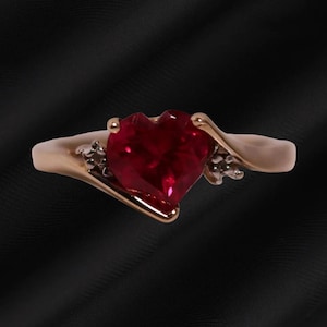 May include: A gold-toned ring featuring a prominent heart-shaped red gemstone at its center. The band has a smooth, flowing design that curves around the stone, accented by small, clear, sparkling stones set on either side of the heart.