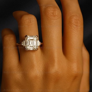May include: A gold ring featuring a large, rectangular, clear gemstone with smaller, rectangular accent stones on each side. The ring is set on a gold band, and the design is displayed against a dark background.