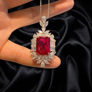 May include: A silver-toned pendant necklace featuring a large, rectangular, vibrant red center stone. The stone is surrounded by a halo of clear, marquise-cut and baguette-cut stones, with a single pear-shaped stone at the top bail.