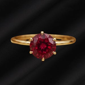 Può includere: Un anello d'oro con una grande gemma di rubino rotonda e sfaccettata. Il rubino è di un colore rosso intenso ed è tenuto in posizione da sei griffe. La fascia dell'anello è in oro lucido.