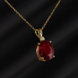 May include: A gold necklace with a large, oval-cut ruby pendant. A small diamond accents the pendant's setting. The chain and pendant setting are gold. The ruby is a deep red color. The background is black.