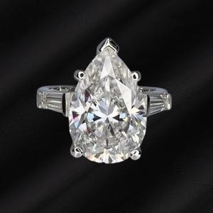 以下が含まれることがあります： 白いゴールドバンドのペアシェイプダイヤモンドリング。リングは、中央に大きな、透明なペアカットダイヤモンドを特徴とし、より小さなバゲットダイヤモンドが両側にあります。バンドには、追加の小さな丸いダイヤモンドがあります。