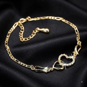 May include: A gold-toned chain bracelet featuring a series of interlocking links and a central focal point consisting of two connected heart shapes. One heart is plain gold, while the other is encrusted with small, clear, sparkling stones.