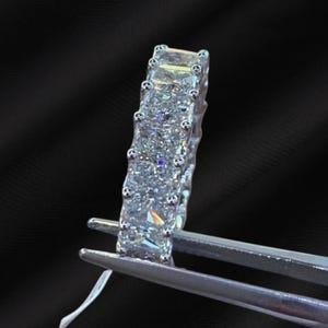 May include: A silver ring with multiple rectangular-cut, clear gemstones. The ring is held by a silver tool against a black background. The gemstones are set closely together, creating a continuous sparkle.