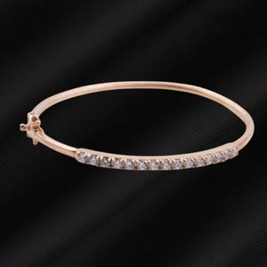 May include: A rose gold bangle bracelet with a row of small, clear gemstones set along the front. The bracelet has a simple, elegant design and a clasp closure. The background is black.