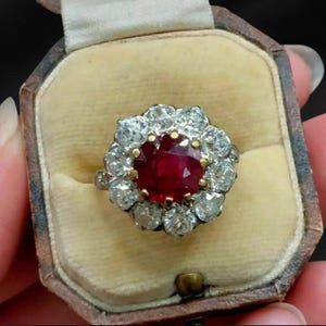 May include: A ring featuring a large, round, deep red ruby at the center, surrounded by a halo of ten sparkling, round-cut white diamonds. The stones are set in a metal band, and the ring is presented inside a vintage-style, octagonal jewelry box.