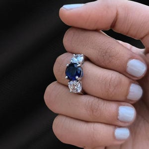 May include: A silver ring with three gemstones: a central dark blue stone, and two smaller, clear stones. The ring is shown on a hand with white nail polish. The gemstones are set in a silver band.
