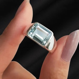May include: A silver ring featuring a large, rectangular, light blue gemstone. The gemstone is set in a polished silver band. The ring is held up against a black background.