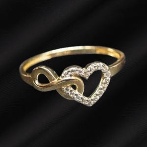 May include: A gold ring featuring an infinity symbol intertwined with a heart shape. The heart portion of the ring is accented with small, clear, sparkling stones set along its perimeter, creating a delicate and continuous metallic band design.