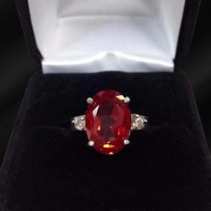 May include: A silver ring with a large, oval-cut, red gemstone as the centerpiece. The ring has smaller, clear stones on either side of the main stone. The ring is displayed in a black jewelry box with a white interior.