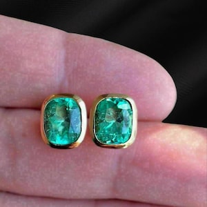 May include: A pair of gold-framed stud earrings, each featuring a vibrant, rectangular emerald-cut green gemstone. The earrings have a polished gold border and a classic, elegant design.