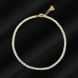 May include: A gold-toned tennis bracelet with a delicate chain and numerous small, round, clear gemstones. The bracelet has a heart-shaped charm at the clasp and is set against a black background.