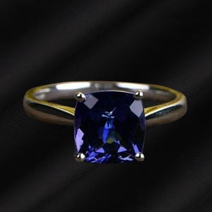 May include: A silver ring with a large, square-cut, deep blue gemstone. The gemstone is set with four prongs, and the ring band is smooth and polished. The background is a dark, textured fabric.