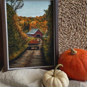 Könnte beinhalten: Ein gerahmtes Gemälde, das eine herbstliche ländliche Szene mit einer roten Scheune und einem mit Kürbissen beladenen Lastwagen auf einem Feldweg zeigt. Die Landschaft besticht durch orangefarbene und gelbe Bäume unter einem hellen Himmel.