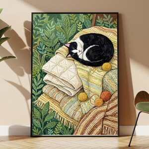 May include: A framed wall art print featuring a black and white cat curled up asleep on a stack of textured blankets and quilts. The scene is surrounded by lush green foliage and includes several small balls of yarn in yellow and orange tones.