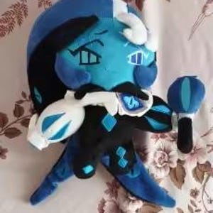 May include: A plush toy character with bright blue skin, black hair, and a blue and white hat. The figure wears a black outfit with blue diamond accents, a white collar, and holds a small blue and black accessory in one hand with a serious expression.