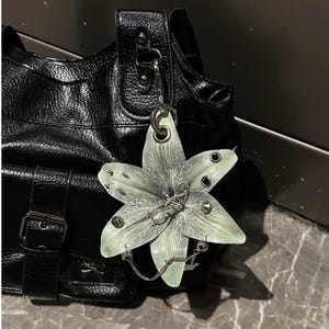 Op de afbeelding: Een zwarte handtas met textuur en een grote, lichtgroene, zesbladige bloemhanger aan de riem. De bloem is versierd met zilverkleurige metalen studs, oogjes en een fijn zilveren kettinkje dat over de onderste bloemblaadjes is gedrapeerd.