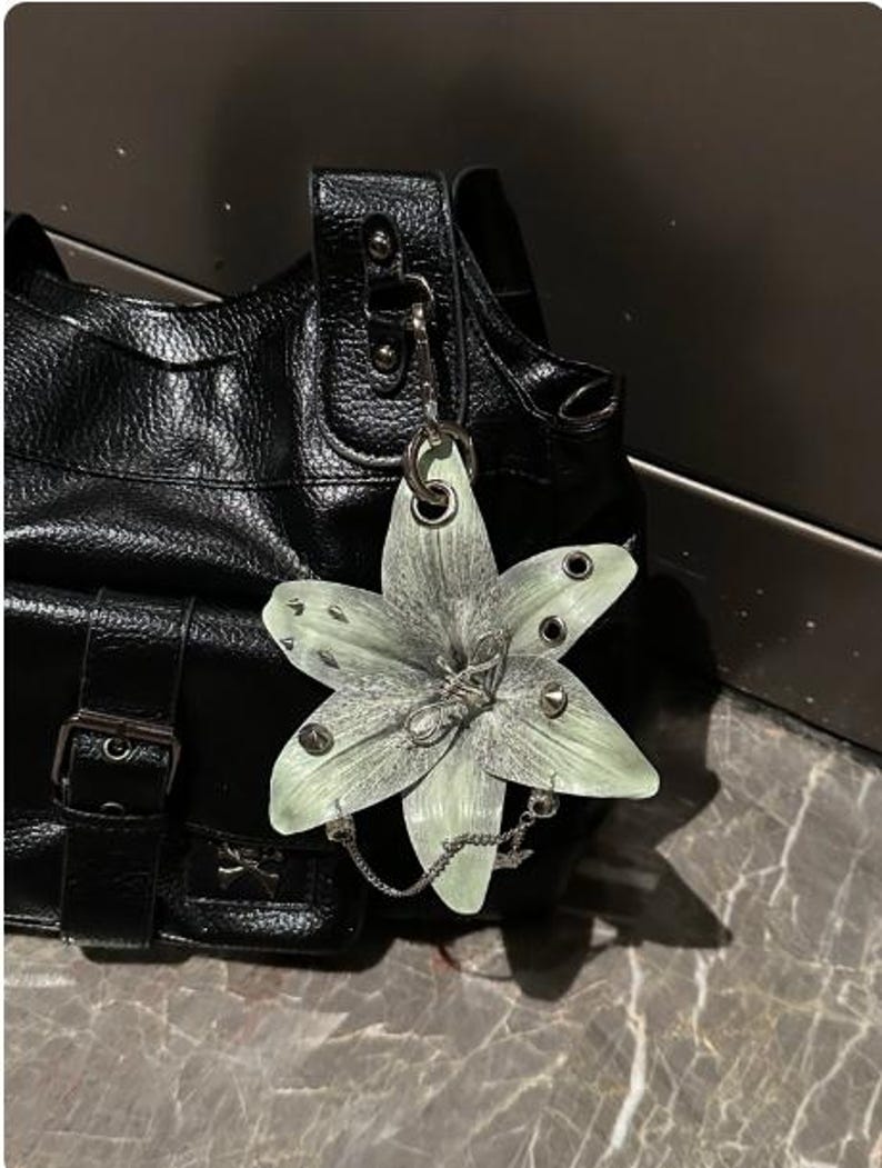 Op de afbeelding: Een zwarte handtas met textuur en een grote, lichtgroene, zesbladige bloemhanger aan de riem. De bloem is versierd met zilverkleurige metalen studs, oogjes en een fijn zilveren kettinkje dat over de onderste bloemblaadjes is gedrapeerd.