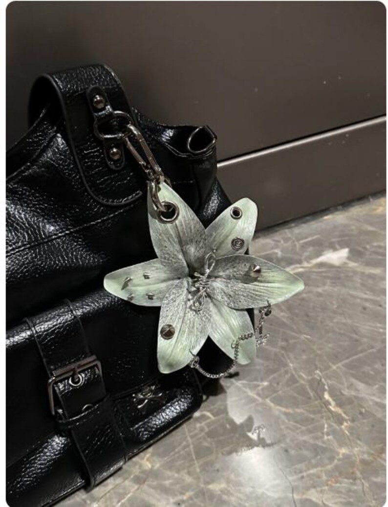 Op de afbeelding: Een zwarte handtas van gestructureerd leer met een zilveren metalen gesp en een grote, lichtgroene lelievormige bedel aan de riem. De bloemversiering is voorzien van kleine zilveren studs, metalen oogjes en een fijn zilveren kettinkje.