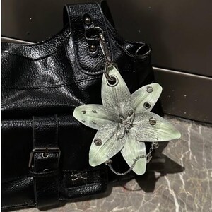Op de afbeelding: Een zwarte handtas van getextureerd leer met een zilverkleurige metalen gespriem en een grote, lichtgroene, zesbladige bloemhanger bevestigd aan een zilveren ketting. De bloem is versierd met kleine zilveren studs en een centraal metalen draaddetail.