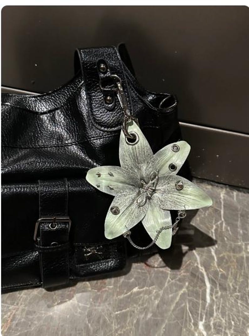 Op de afbeelding: Een zwarte handtas van getextureerd leer met een zilverkleurige metalen gespriem en een grote, lichtgroene, zesbladige bloemhanger bevestigd aan een zilveren ketting. De bloem is versierd met kleine zilveren studs en een centraal metalen draaddetail.