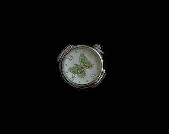 Op de afbeelding: Een zilverkleurige ronde wijzerplaat met een witte achtergrond en een groene vlindergrafiek in het midden. De wijzerplaat toont zwarte Romeinse cijfers op de posities drie, zes, negen en twaalf uur, met dunne goudkleurige wijzers.
