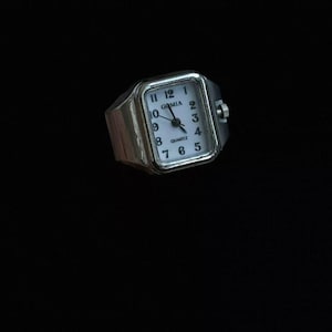 May include: A silver-toned ring featuring a rectangular watch face with a white dial, black Arabic numerals, and the text GOMIA and QUARTZ printed on the face. The watch has a small crown on the right side and black hour, minute, and second hands.