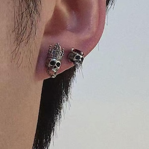 May include: Two silver-toned stud earrings worn on an earlobe. One earring features a detailed skull wearing a crown, while the other earring is a smaller skull design topped with a dark red gemstone. Both pieces have an oxidized finish.