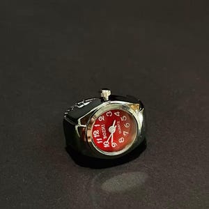 May include: A silver-toned metal ring watch featuring a round face with a vibrant red dial. The watch face displays white Arabic numerals from 1 to 12, the brand name BOZHI, and the word QUARTZ. A small silver crown is positioned at the top.